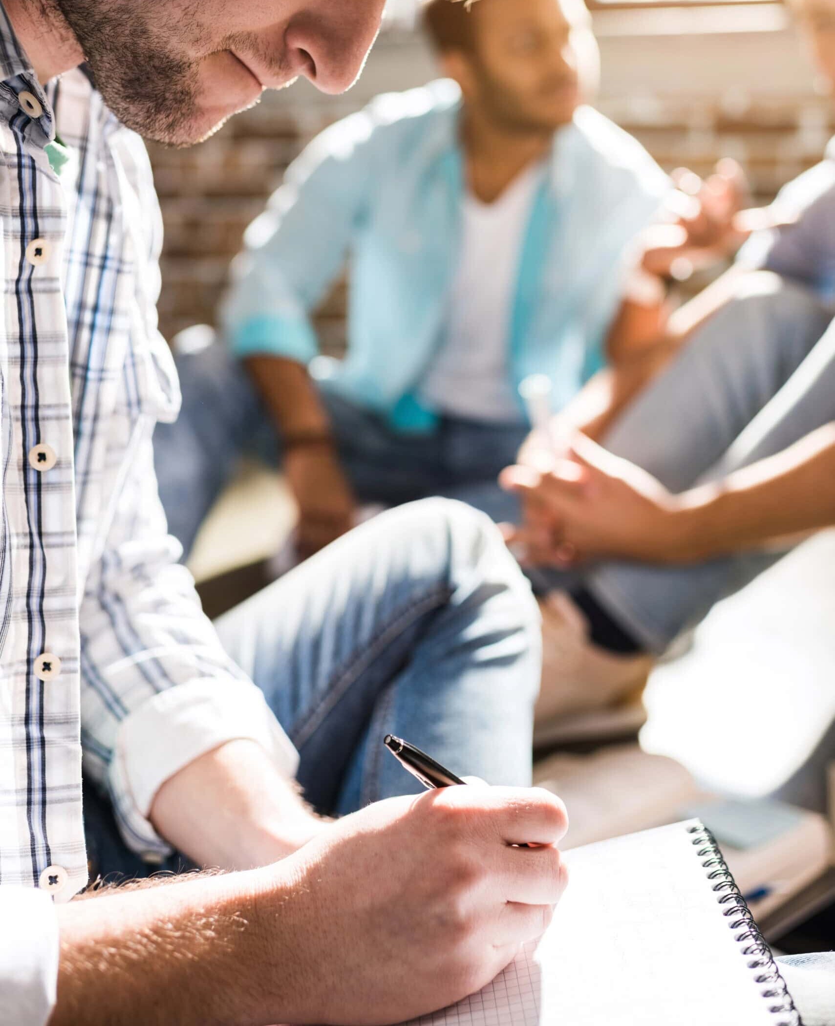 a focused young man writes in a notebook, capturing ideas during a group discussion